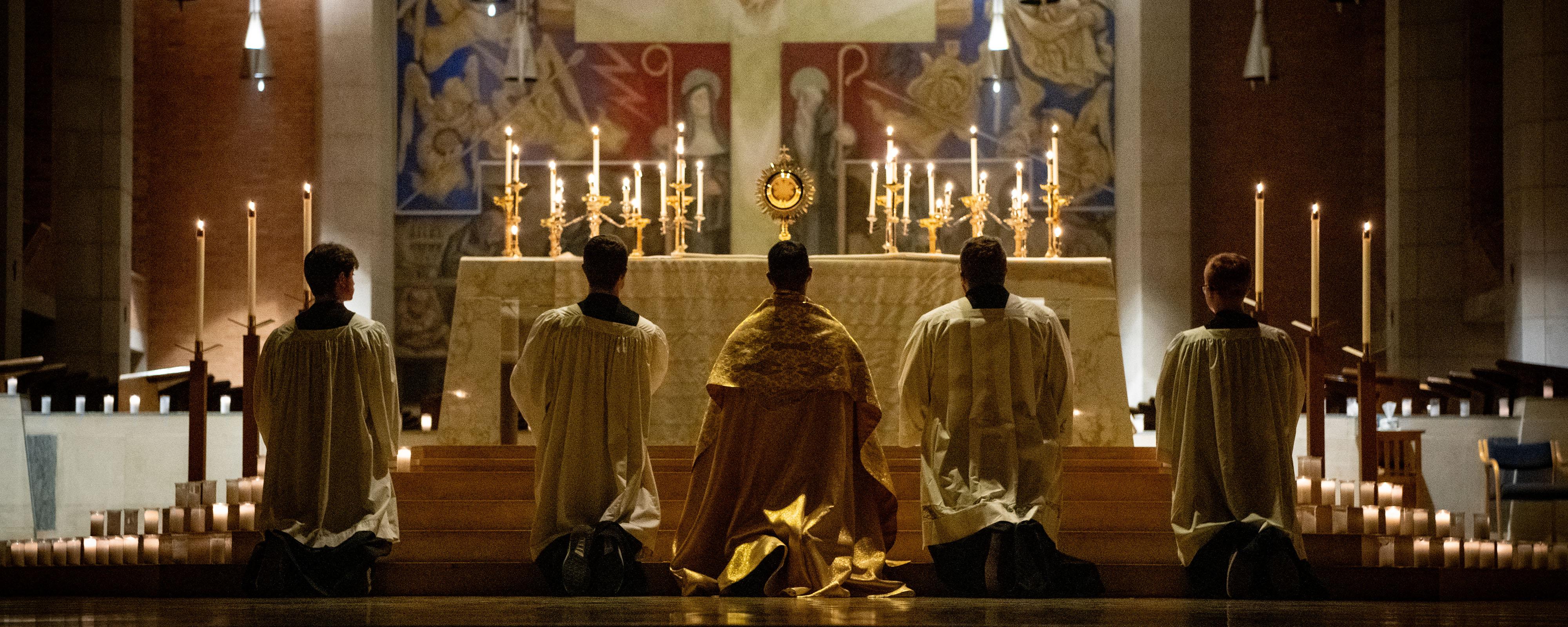 Priest and 4 altar servers kneeling in adoration with votive candles